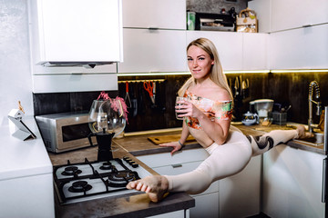 Flexible girl is drinking coffee. Young gymnast doing the splits on the table. Yoga, sport stretching in the kitchen