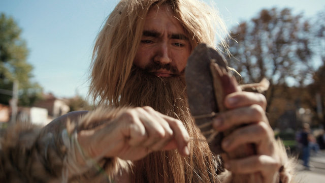 Close-up Of Neanderthal Savage Making A Stone-tipped Spear Creating Hunting Tool Staying In Modern City Street. Wild Homo Sapiens. Evolution.