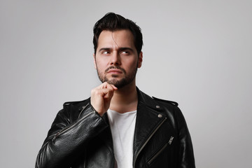 Shot of brutal man with dark beard, looks aside, wears leather jacket, over blank studio background with free space.