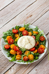 Tasty, fresh salad with mozzarella burrata, arugula, cherry tomatoes and basil on a white plate on a wooden background.Close up. Healthy Italian food. Selective focus