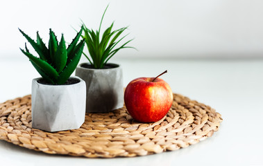 Cozy home interior decor: red apple and ornamental plants in pots on a wicker stand on a white table in the room. The quarantine concept of stay home.
