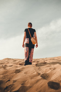Young woman (adventurer) in oriental clothes walking through the desert - photo from back. Footsteps in the sand of walking woman (traveler and turist) with a bag on desert - moody shot.