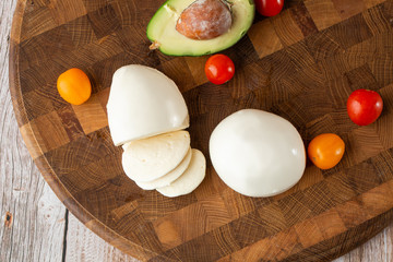 Overhead photo of buffalo mozzarella cheese with cherry tomatoes, basil leaves, avocado, ingredients of salad, pesto sauce, olive oil, on rustic wooden cutting board with copy space. Italian cheese