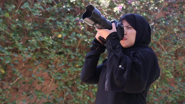 A beautiful Moroccan Arab Muslim woman with a DSLR camera filming photographing in a garden - turning the camera up and towards the viewer. Real-time footage.