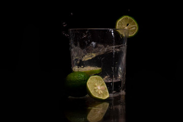 Water splashes out of the garnished glass as a green fresh lemon slice immerses into the water.