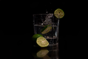 Water splashes out of the garnished glass as a green fresh lemon slice immerses into the water.