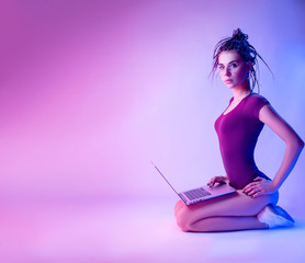 a girl dressed in a bodysuit with dreadlocks on her head is sitting on the floor on a white background in neon light to looking at a laptop