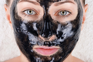 A beautiful girl with a black mask on her face is lying on a white soft towel very close-up. Black face mask with activated carbon. Face care in the salon or at home, cleaning the pores.