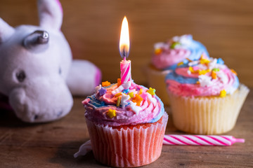 Unicorn cupcakes with a lit candle decorated with pink icing and sprinkles on a dark wooden background