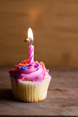 Single cupcake with a lit candle decorated with pink icing and sprinkles on a dark wooden background