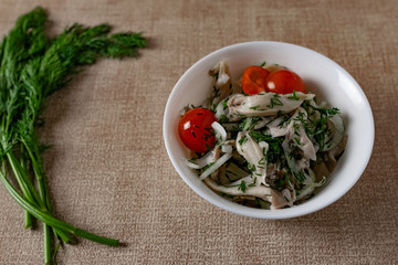 Marinated oyster mushrooms, garnished with cherry tomatoes and green dill. A dish with onions and butter in a white plate with a beige light cloth on the table