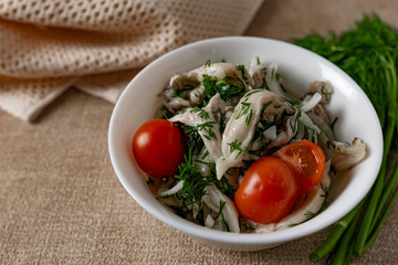 Marinated oyster mushrooms, garnished with cherry tomatoes and green dill. A dish with onions and butter in a white plate with a beige light cloth on the table