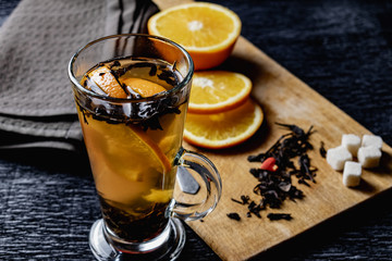Citrus drink, tea with petals in a transparent glass mug with slices of sweet orange and refined sugar. Slicing fruit on a wooden Board with a brown napkin in a cozy dark background in a low key