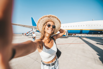 Beautiful happy woman taking a selfie at the airport in front of the plane. People departure boarding holidays concept.  - obrazy, fototapety, plakaty