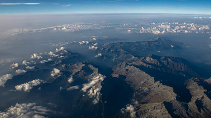 Beautiful aerial view on the Italian Alps on a sunny day with clear sky and low altitude clouds. Flying above mountains with some haze in background