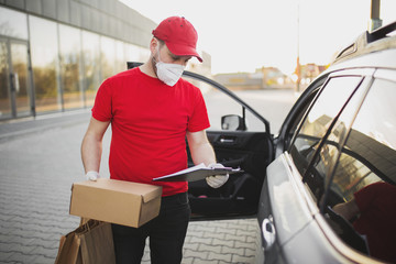 Deliveryman in red uniform with brown paper package in medical respirator in vehicle