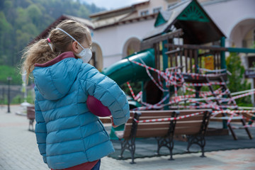 Blond hair girl, wearing a medical mask, closed playground on the background. A child in a...