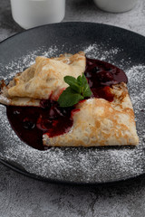 Crepes with cherry jam folded in triangles decorated with powdered sugar, on black plate, close mouthwatering shot