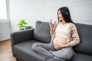 Beautiful asian pregnant woman drinking holding glass of milk, eating healthy having lunch sitting on the grey sofa, smiling joyfully relaxing and resting living room with white brick wall texture