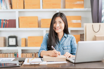 beautiful Asian businesswoman smiling using writing on parcel packaging, using computer laptop ecommerce online store selling delivering products, working at home office during quarantines isolation