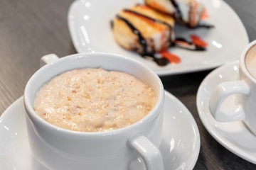 Oatmeal porridge on a wooden table near cheesecakes and cappuccino.