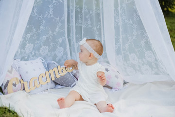 A little girl is sitting under a canopy on a white blanket. Cute girl in profile in a white dress and headband in nature. Baby girl holding a sign " Happiness" © Яна Айбазова