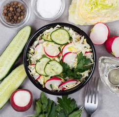 Vitamin salad of fresh vegetables cabbage, radishes, cucumbers and parsley in a bowl on a light gray background top view