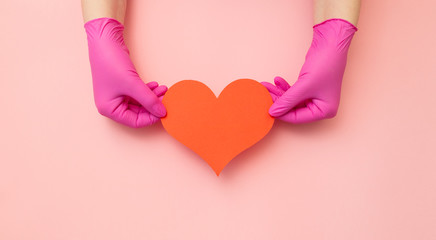 Sign of heart keeping female hands in medical red gloves on pink background. Banner with copy space...
