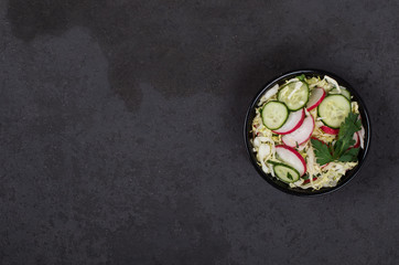 Vegan salad with fresh cabbage, radish, cucumber and parsley in a bowl on a dark background top view