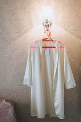 Female white bathrobe, underwear hanging on a red hanger against the background of a wall and a night lamp. Evening bedtime. Photography, concept.