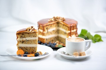a cup of coffee with two pieces of sugar on a saucer and next to it a piece of carrot cake on a white plate, covered with caramel and decorated with nuts on top, in the background is a cake and a spri