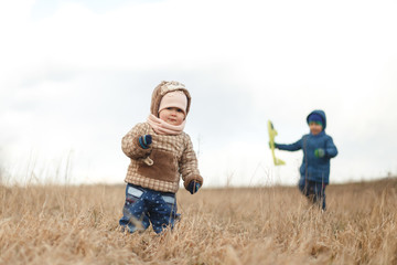 Naklejka na ściany i meble Little baby girl standing in the field of dry grass and her brother with toy airplane. Portrait with background of white sky.