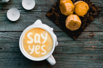 Black coffee with milk foam with the inscription pattern say yes in a white cup on an old painted and peeling wooden background. Boiled condensed rolls surrounded by candles