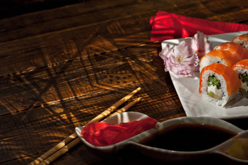 Sushi on a white plate. Sushi roll with sauce and spices on a black background. Food on a wooden table with dark boards. Sakura color on a plate. Flower with rose petals. Place for text near land.