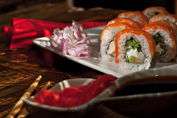 Sushi on a white plate. Sushi roll with sauce and spices on a black background. Food on a wooden table with dark boards. Sakura color on a plate. Flower with rose petals.