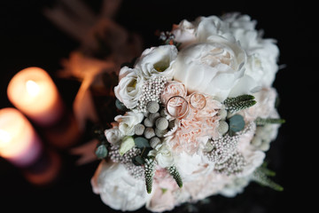 Wedding bouquet of white, pink and green flowers with gold wedding rings near burning candles on a black background