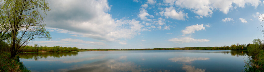 Panoramic landscape from the lake shore with colorful clouds in the spring sun.