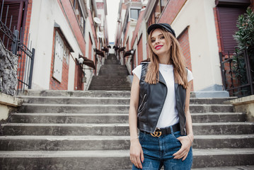 Beautiful in a leather vest, jeans with stars and a cap stands in the city on the stairs