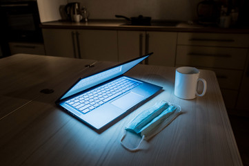 Remote work overtime during the quarantine period in the home office. Working atmosphere in the kitchen on a laptop in the dark late at night. Coffee mug and medical mask on the table.