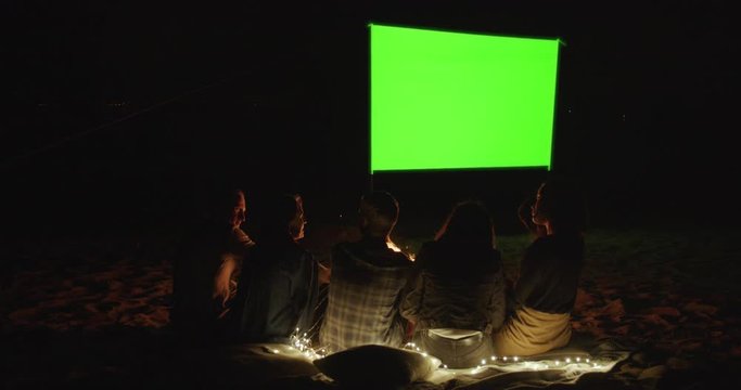 Outdoor cinema. Friends gather in a campfire camp and watch a movie with a projector on the beach near the sea. Green screen. A place for your image