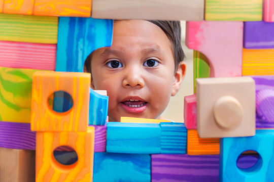 A young boy showing his face through a window from behind a wall of building blocks with various shapes and colors.