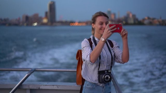 Happy female blogger in casual clothing using cellphone camera for clicking pictures of evening metropolitan cityscape during water boat excursion, cheerful hipster girl photographing via cellular
