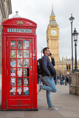 London, UK - April 2, 2017: Casual man talking on mobile phone in London, leaning on traditional british red telephone booth. Big Ben can be seen in background on 2th of April, 2017 in London. - obrazy, fototapety, plakaty