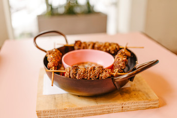 Wooden container with skewered meat served with a bowl of sauce in the middle on a wooden table