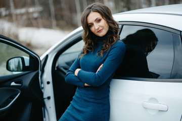 Young beautiful girl stands in a dress by a white car. Сoncept of car breakdown on the road. Brunette is waiting for help on the road