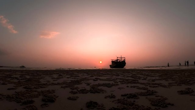 Sun goes down near the ship while people walk over sandy beach timelapse sunset. Concept of vacation at the beach and travelling. Timelapse of the sunset behind the standing boat at the seashore.