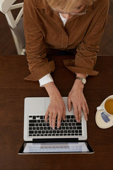 Female With Types On Laptop. Work From Home Concept. Blonde Woman in Brown Jacket And Eyeglasses Uses Notebook On Desk.