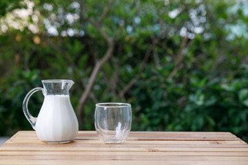 jug of milk and cup on a wooden table at green tree background