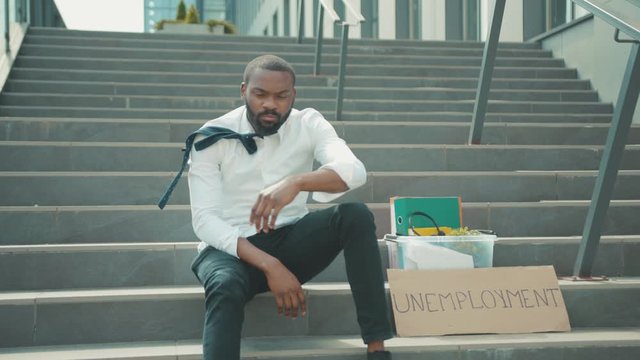 Sad African american young man sitting on stairs use phone with box personal stuff and poster with the inscription Unemployment got fired due to coronavirus crisis depression economy slow motion