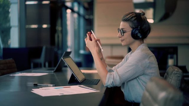Happy female student in joyful mood choosing music songs from smartphone podcast for listening via electronic bluetooth headphones during elearning on digital tablet, Caucasian woman working remotely
